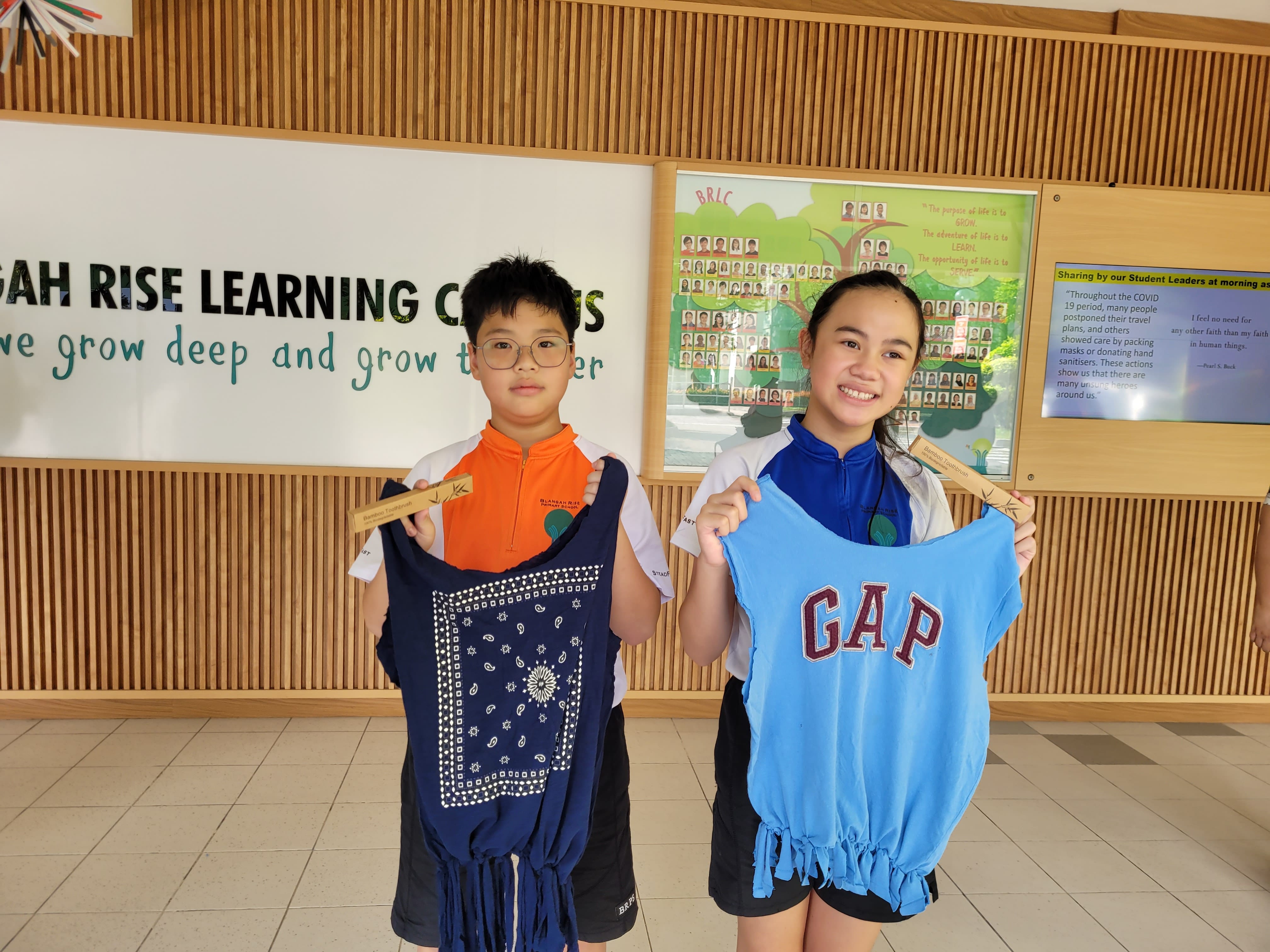 Two children hold upcycled fringed shirts and bamboo toothbrushes inside a school learning center.